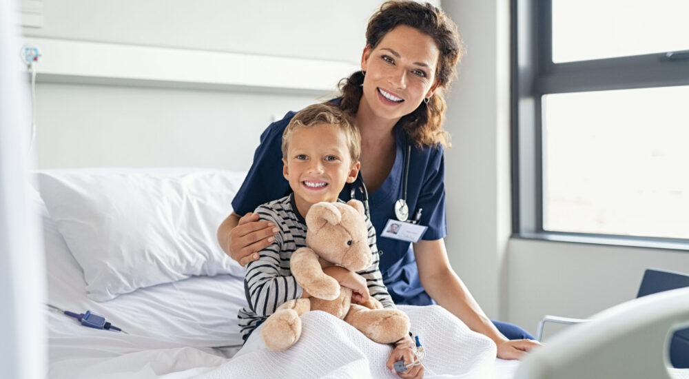 Portrait of cheerful little boy holding teddy bear while sitting with beautiful nurse on gurney at hospital. Happy female nurse in pediatric ward with kid looking at camera. Young pediatrician doctor visiting and comforting hospitalized child at private clinic.