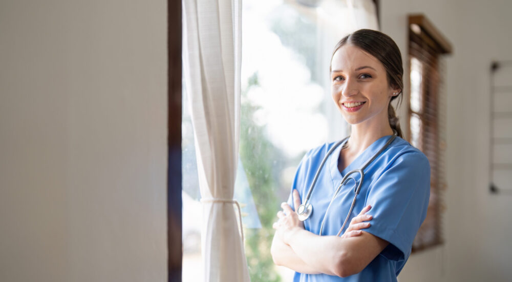 Portrait of a friendly female doctor or nurse wearing blue scrubs uniform and stethoscope, with arms crossed in hospital.